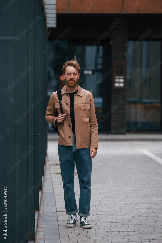 Front view. Stylish man with beard in khaki colored jacket and in jeans is outdoors near building