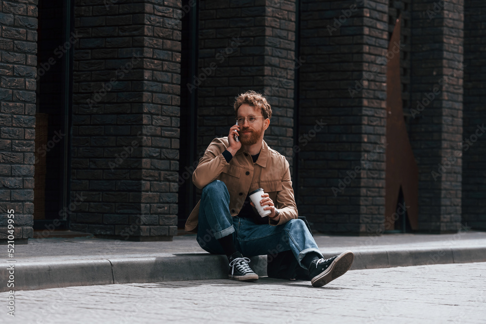 With phone. Stylish man with beard in khaki colored jacket and in jeans is outdoors near building