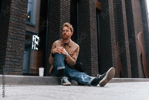 Coffee cup. Stylish man with beard in khaki colored jacket and in jeans is outdoors near building