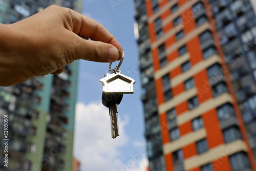 Real estate agent, keychain in shape of a house and key in male hand on background of new buildings. Byung apartment or renting property in summer