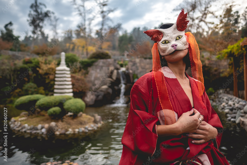 Portrait of sexy and young japanese girl with beautiful old traditional red kimono and handmade cat mask in front of a pond in a japanese garden