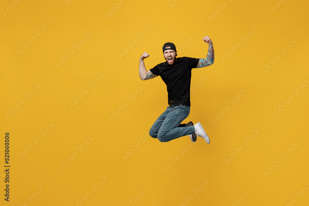 Full body young overjoyed cool bearded tattooed man 20s he wears casual black t-shirt cap jump high do winer gesture isolated on plain yellow wall background studio portrait. People lifestyle concept.