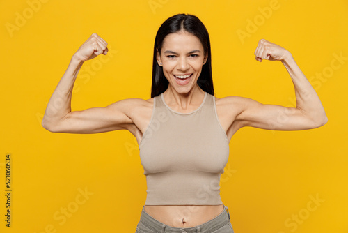 Young strong sporty fitness latin woman 30s she wear basic beige tank shirt showing biceps muscles on hand demonstrating strength power isolated on plain yellow backround. People lifestyle concept