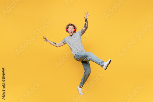 Full body young man 20s he wear grey t-shirt headphones listen to music stand on toes with outstretched hands raise up leg isolated on plain yellow backround studio portrait. People lifestyle concept. © ViDi Studio