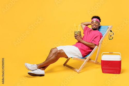 Full size young man he wear pink t-shirt bandana lying on deckchair near hotel pool drink pineapple juice hold hand behind neck isolated on plain yellow background. Summer vacation sea rest concept.