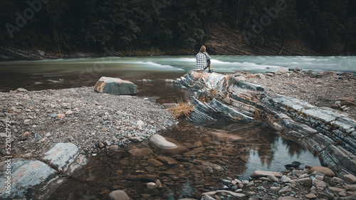 A man with a beard in a plaid shirt and shorts sits on the bank of a mountain river, moody colors