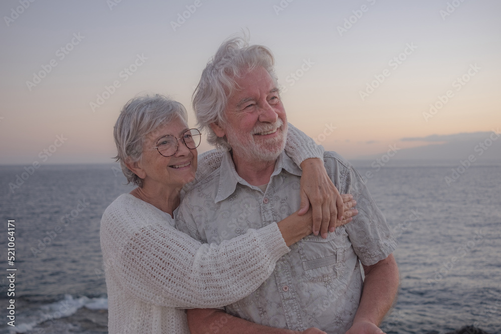Portrait of two happy and romantic seniors or pensioners embraced at the sea at sunset light - old smiling senior couple outdoors enjoying vacations together