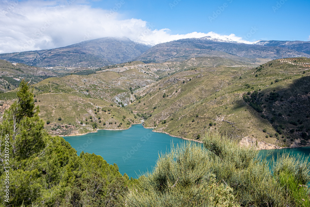 View from the top on green lake surrounded by picturesque mountain alley with trees and bushes with blue sky and white clouds in background in sunny summer day.