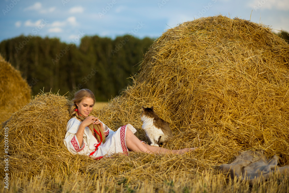 Slavic woman in national costume sitting in a haystack in a field braids her hair. Cat in the hay.