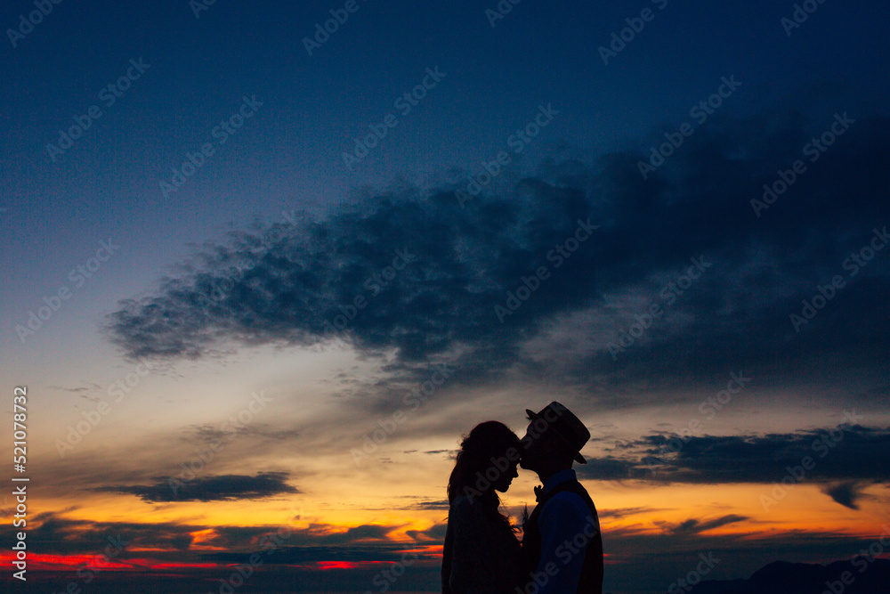 Silhouette of groom in the hat kisses bride on the forehead at sunset