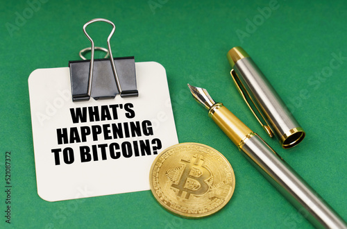 On a green surface, a bitcoin coin, a pen and a sheet of paper with the inscription - What is happening to Bitcoin