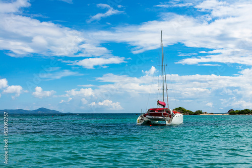 white catamaran with a red sail near a tropical island against a blue sky with white clouds. Tropical paradise concept