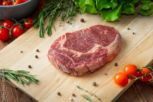 Raw Ribeye steak or beef steak on a wooden cutting board with tomatoes and herbs around