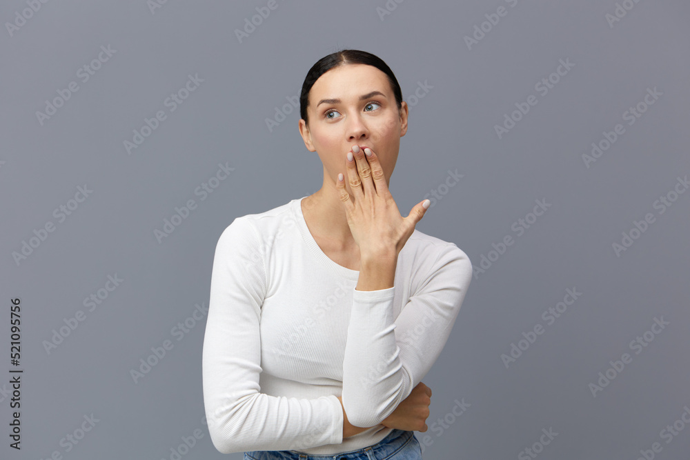 a cute woman with a ponytail on her head stands on a dark background in a white tight T-shirt, covered her mouth with her hand in surprise a cute woman with a ponytail on her head stands on a dark background in a white tight T-shirt, covered her mouth with her hand in surprise