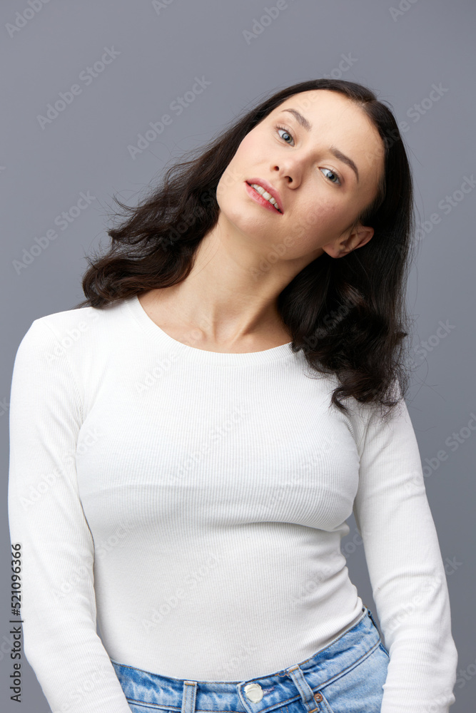 a sophisticated woman stands on a gray background in a white T-shirt, tilted her head to the side, lowering her hands down, smiling gently