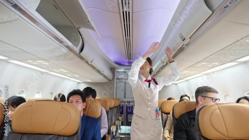 Asian woman flight attendant walking on airplane aisle closing luggage compartment over passenger seat before aircraft take off runway. Cabin crew checking safety and passenger security in the plane photo