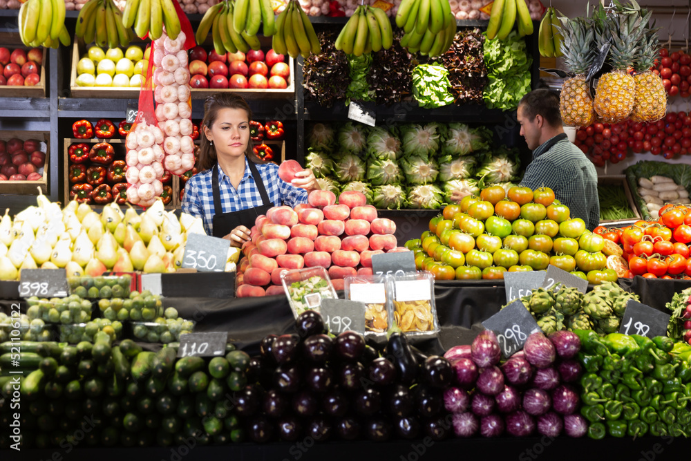 Man and woman shop assistants laying out fruits and vegetables behind the counter on market