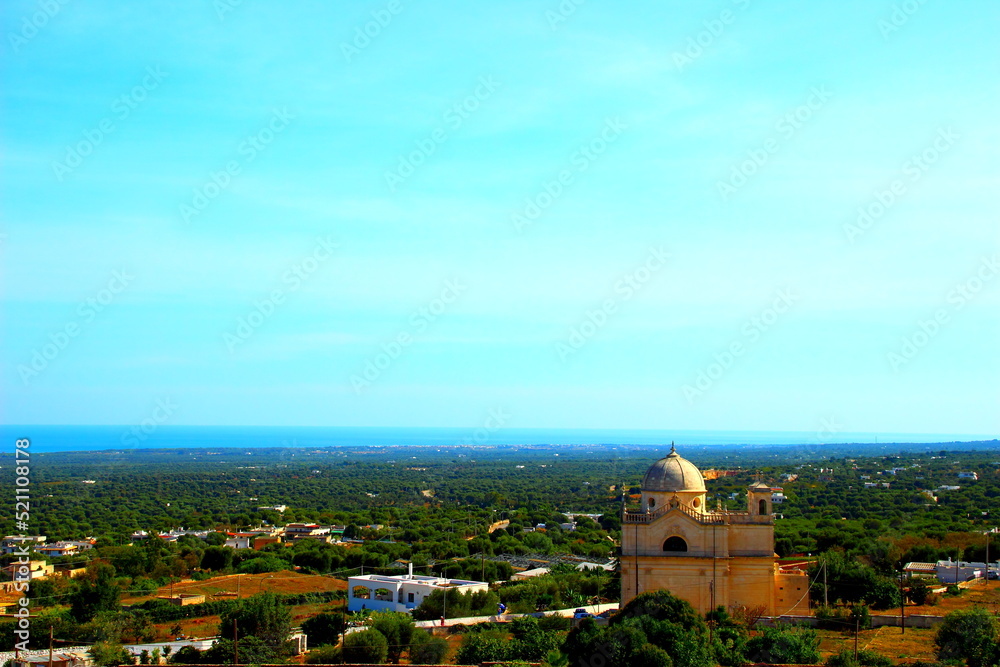 Fabulous overview from a hill in Ostuni with a church and sporadic white houses bathed in lush deep green vegetation and the magnificent hazy colours of the Adriatic Sea in the background