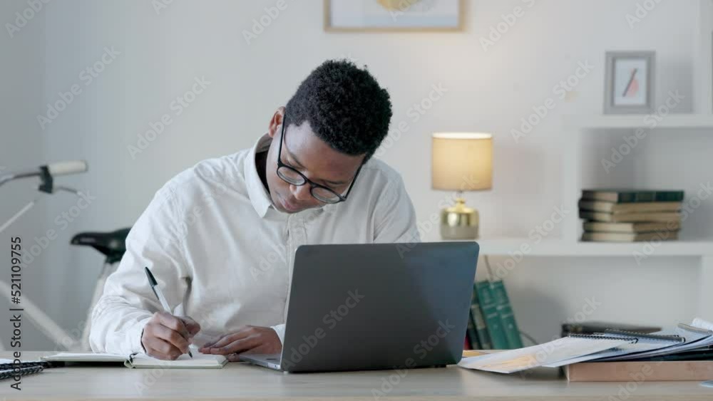Black business man writing notes while working remotely on laptop, analyzing documents in a home office. African American financial analyst checking documents while making a list for a finance report Black business man writing notes while working remotely on laptop, analyzing documents in a home office. African American financial analyst checking documents while making a list for a finance report