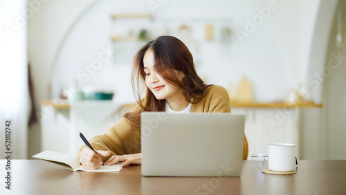 Young asian woman sitting at table and taking notes in notebook. Working on table with computer laptop, document paperwork and cup of coffee. Student learning online