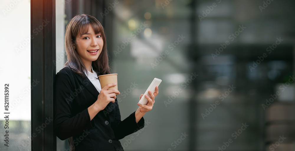Portrait of young businesswoman holding coffee cup and using mobile phone, standing at office, smiling and looking at camera. Portrait of young businesswoman holding coffee cup and using mobile phone, standing at office, smiling and looking at camera.