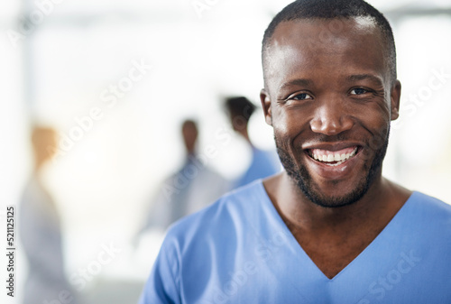 Happy  smiling and professional doctor in a hospital closeup portrait with blurred background. Confident black male medical healthcare worker with colleagues in the back. An African American surgeon.