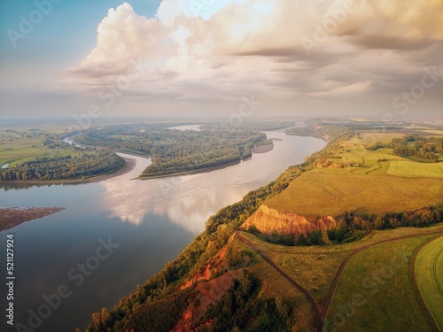 In large clouds, the light of the setting sun and the warm evening light flood the trees on the hill, the clay mountain and the reflection of the clouds in the water. river Ob.