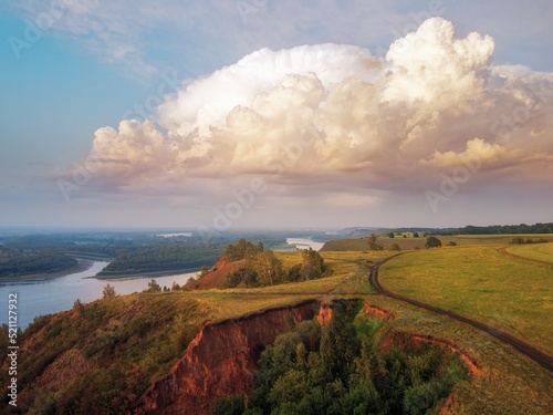 In large clouds, the light of the setting sun and the warm evening light flood the trees on the hill, the clay mountain and the reflection of the clouds in the water. river Ob.