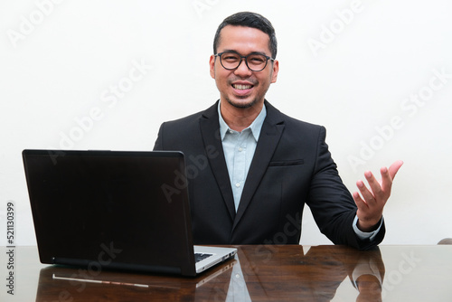 Asian man wearing black suit smiling friendly in front of his laptop