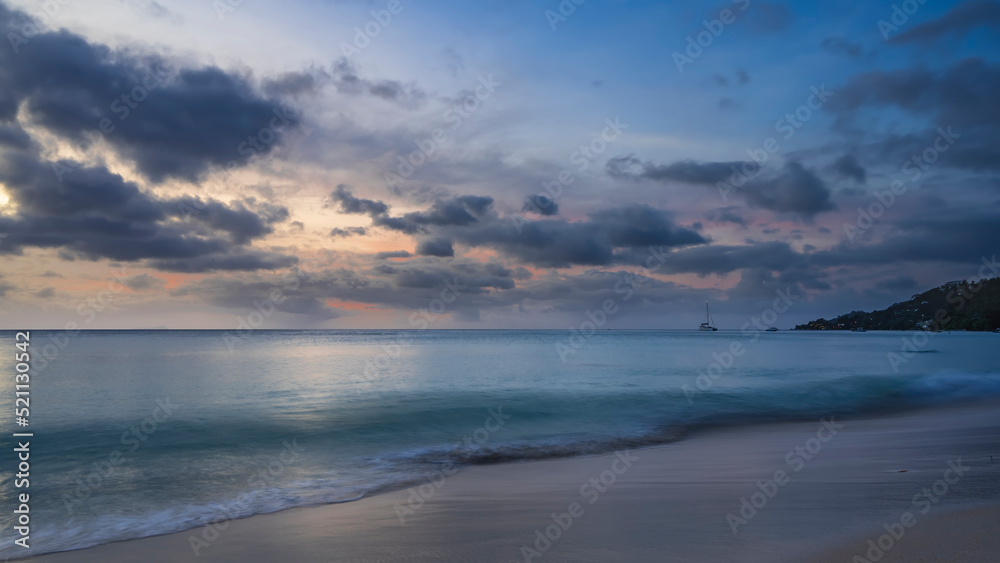 Blue clouds in the evening sky, highlighted in pink and orange. Reflection on the surface of the turquoise ocean. Foam of waves on a sandy beach. Long exposure. Seychelles. Mahe. Beau Vallon beach