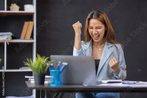 Happy excited successful Asian businesswoman triumphing with a laptop computer smartphone in the workplace office