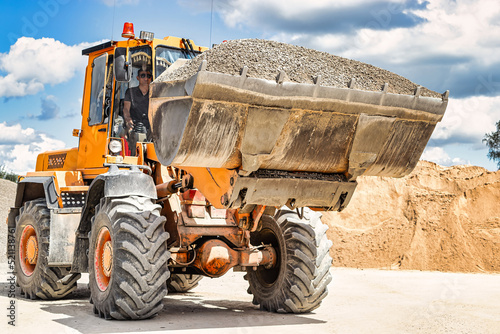 A large front loader transports crushed stone or gravel in a bucket at a construction site or concrete plant. Transportation of bulk materials. Construction equipment. Bulk cargo transportation.