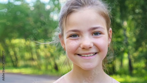 Schoolgirl with green eyes stands on blurry green forest pathway. Happy girl looks straight smiling. Teen girl enjoys walking in forest closeup