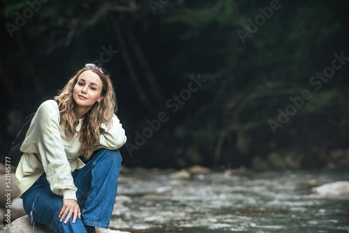 portrait of a beautiful traveler girl with long wavy hair against the backdrop of a green forest and a mountain river