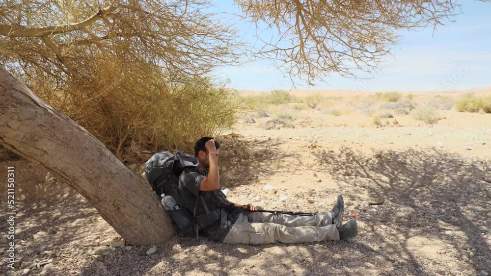 Caucasian hiker backpacker with trekking poles rest sitting under dry tree during travel in Makhtesh Crater Ramon, in the Negev Desert, Southern Israel. Caucasian hiker backpacker with trekking poles rest sitting under dry tree during travel in Makhtesh Crater Ramon, in the Negev Desert, Southern Israel.