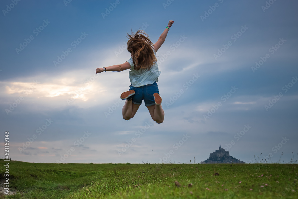 Young, blonde girl taking a big leap, with Mont Saint-Michel in the background. France Young, blonde girl taking a big leap, with Mont Saint-Michel in the background. France