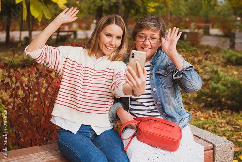 charming women , mom and daughter are sitting on a bench in a beautiful autumn park and video chat
