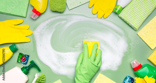 Home cleaning products. A hand in a green rubber glove wipes soap suds with a washcloth on a green background. Concept of cleanliness, cleaning services.