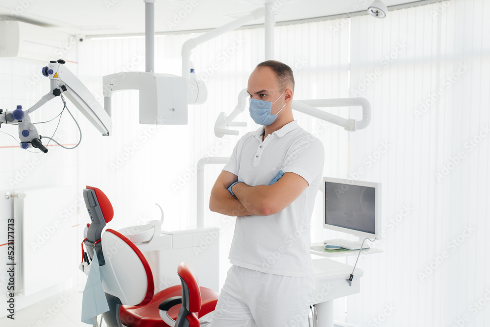 Obraz premium A young dentist in a mask stands near a red dental chair and smiles in modern white dentistry. Modern dentistry and prosthetics.