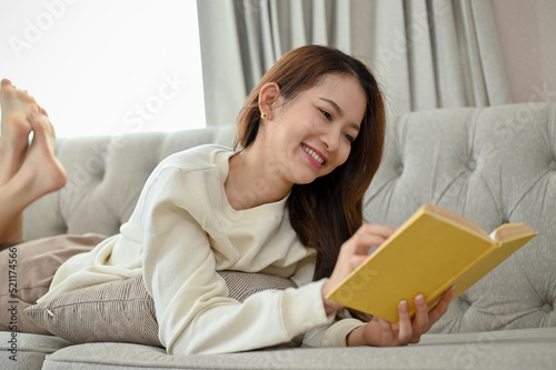 Attractive Asian female laying on a sofa in the living room, enjoy reading an adventure fiction book