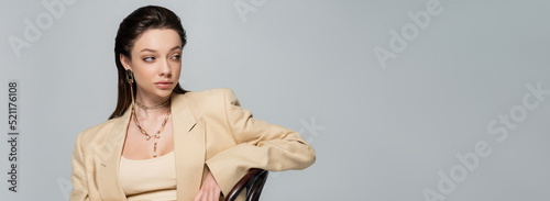 brunette young woman in stylish outfit looking away and leaning on chair isolated on grey, banner