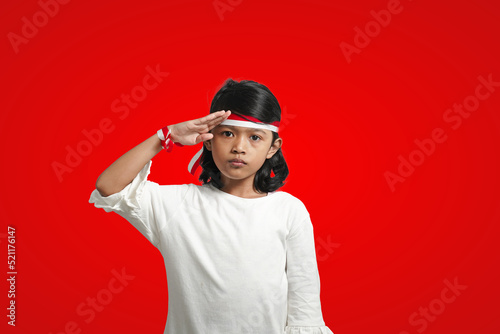 Isolated girl wearing red and white ribbon celebrates Indonesia's independence by saluting in front of the camera.