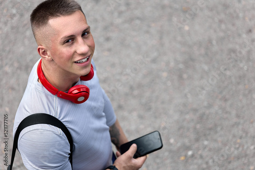 View from above of a smiling young athlete with a phone in his hand and red headphones heading to training. A happy man with a smartphone smiles, looks at the camera with his head up photo