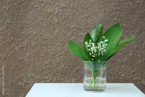 Beautiful lily of the valley flowers in glass vase on white table near brown wall  space for text