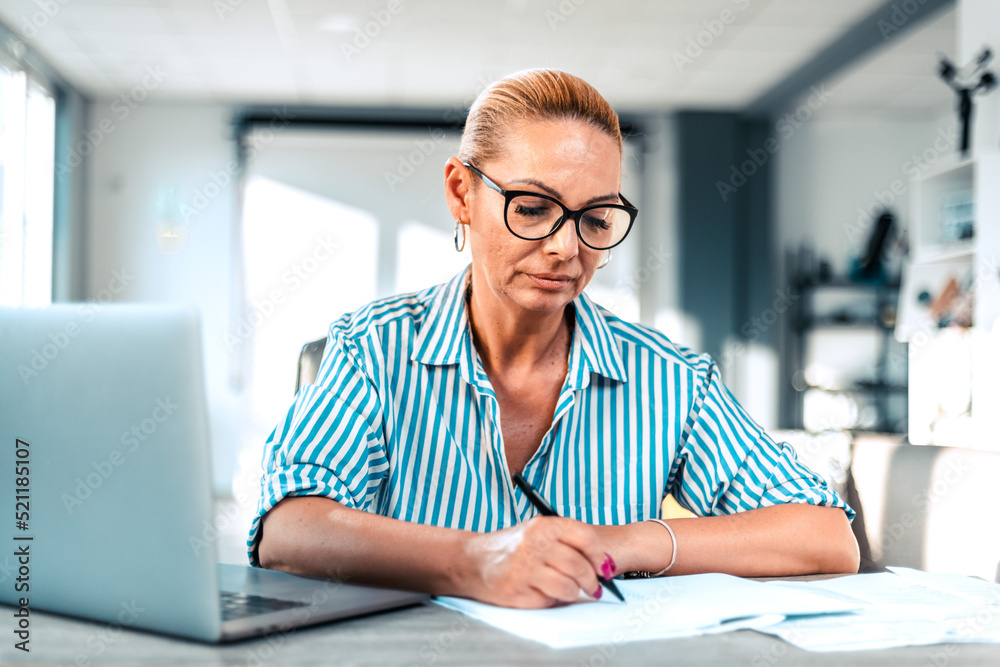Portrait of confident mature female accountant writing on documents at desk in office. Portrait of confident mature female accountant writing on documents at desk in office.