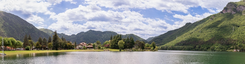 Super widescreen shot of gently sloping shore of a high-altitude glacial lake Lago d'Idro, evergreen trees under dazzling white clouds. Brescia, Lombardy, Italy