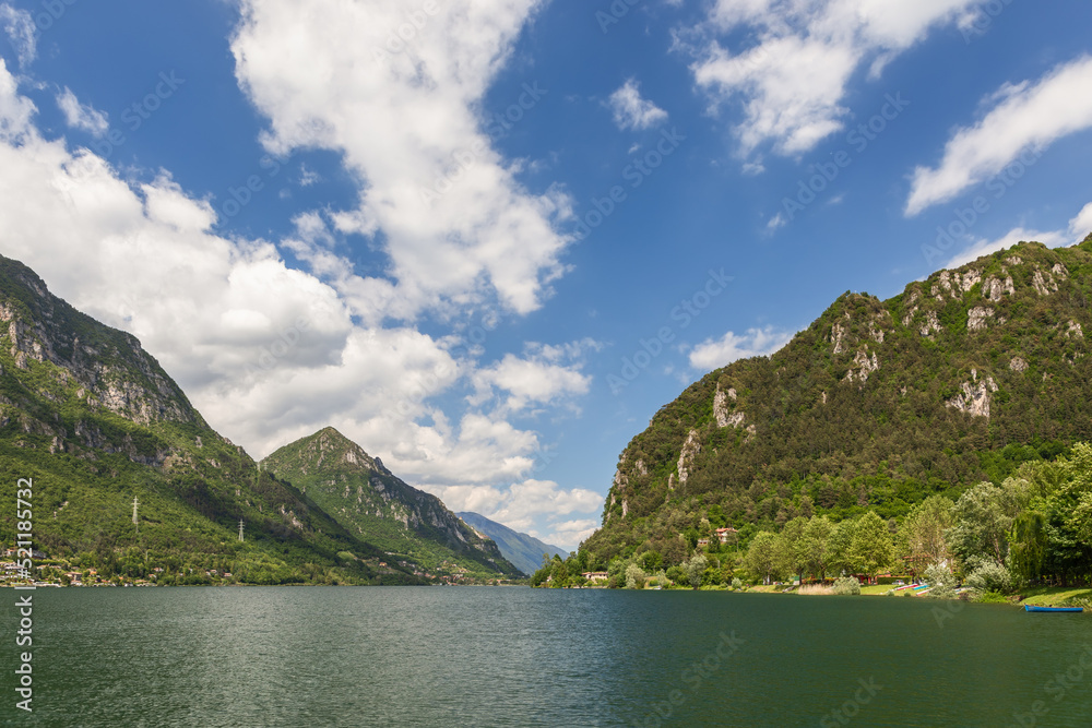 Green waters of the lake (Lago d'Idro) framed by alpine rocks with evergreen trees under a clear blue sky with white clouds, wide panorama. Brescia, Lombardy, Italy