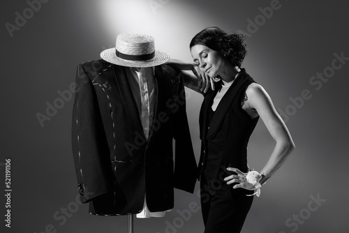 Portrait of stylish woman, fashion designer in classic black outfit leaning on mannequin in male costume. Black and white photography