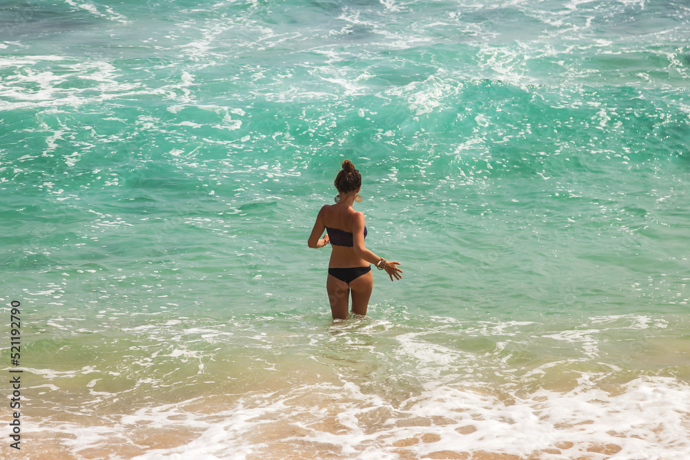 Back view young woman with slim body in tropical ocean wearing bikini in waves