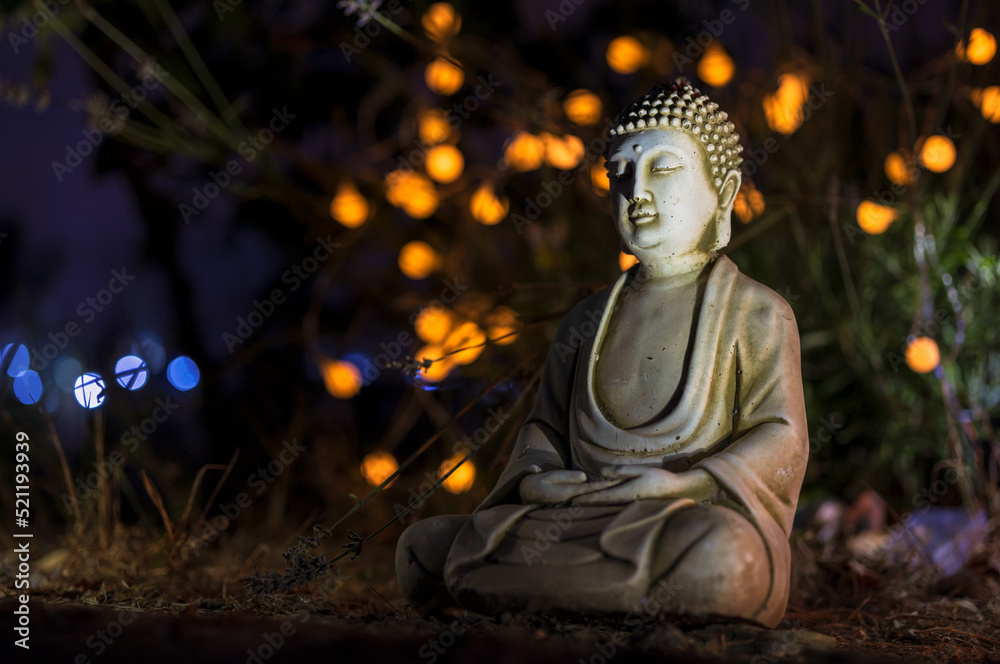 Figure of a Buddha with joined hands in a natural garden with small lights.
